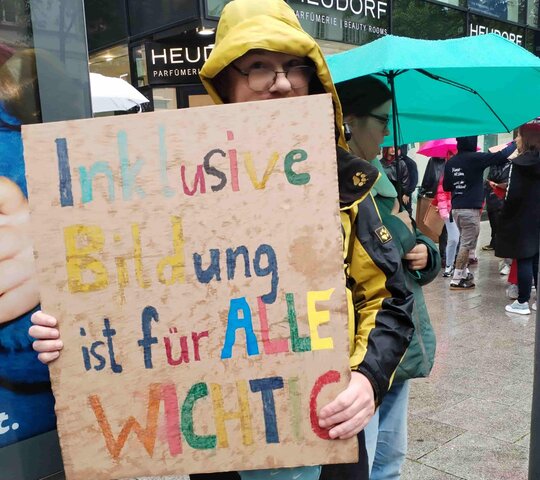 Bildungsfachkraft Thilo Krahnke auf der Kundgebung in Stuttgart. Er trägt ein selbst gestaltetes Plakat vor sich. Auf dem Plakat steht in bunten Buchstaben: Inklusive Bildung ist für alle wichtig.