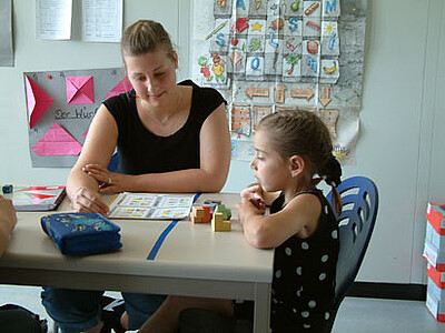 Auf dem Bild sieht man eine typische Fördersituation. Eine Studentin sitzt mit einem Kind an einem Tisch und sehen sich geometrische Körper an.