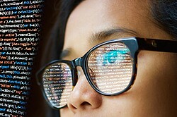 Das Foto zeigt technische Codes, die sich n der Brille einer Frau spiegeln.