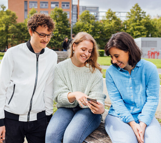 Drei Personen sitzen auf einer Mauer und schauen gemeinsam in ein Handy