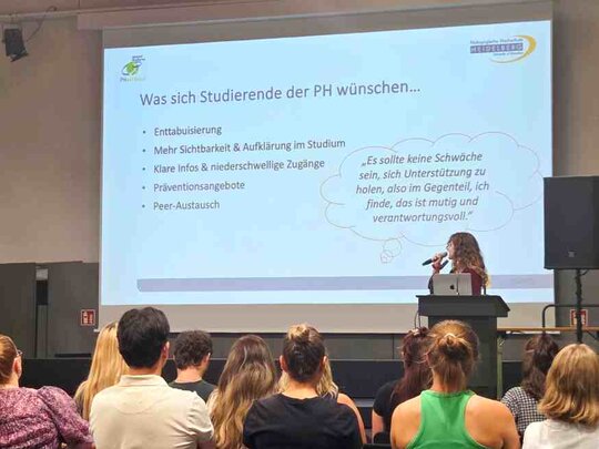 Eine Studentin steht auf der Bühne in der Festhalle. Man sieht eine Präsentationsfolie. Im Publikum sitzen zahlreiche Personen, die man von hinten sieht.