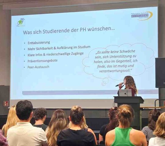 Eine Studentin steht auf der Bühne in der Festhalle. Man sieht eine Präsentationsfolie. Im Publikum sitzen zahlreiche Personen, die man von hinten sieht.
