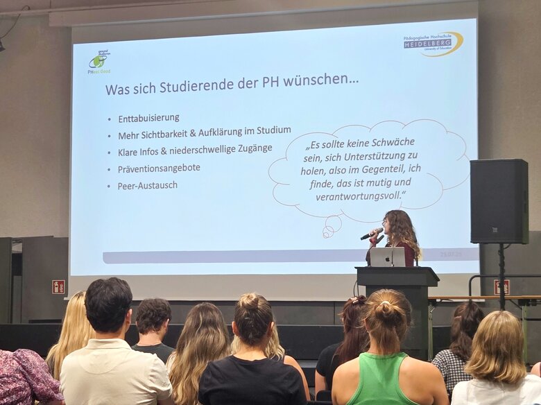 Eine Studentin steht auf der Bühne in der Festhalle. Man sieht eine Präsentationsfolie. Im Publikum sitzen zahlreiche Personen, die man von hinten sieht.