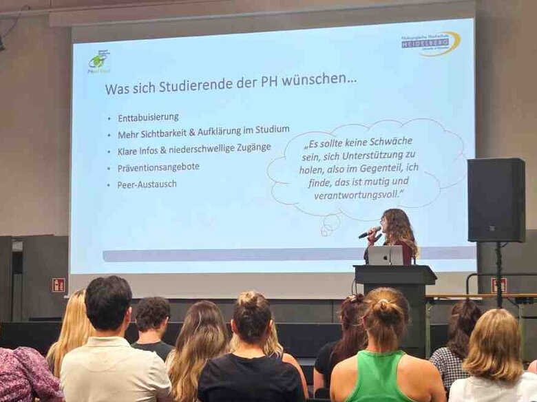 Eine Studentin steht auf der Bühne in der Festhalle. Man sieht eine Präsentationsfolie. Im Publikum sitzen zahlreiche Personen, die man von hinten sieht.