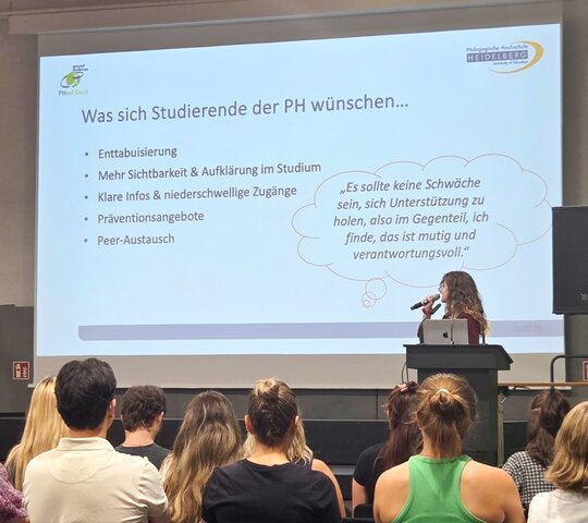 Eine Studentin steht auf der Bühne in der Festhalle. Man sieht eine Präsentationsfolie. Im Publikum sitzen zahlreiche Personen, die man von hinten sieht.