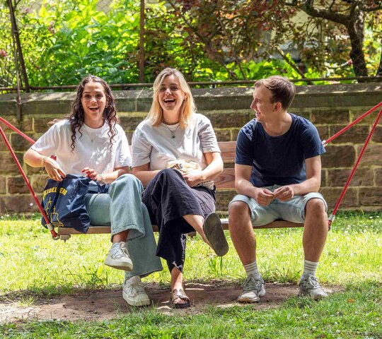 Zwei Studentinnen und ein Student sitzen in der Hängeschaukel. Alle drei lachen deutlich. Im Hintergrund sieht man den Innenhof der Hochschule.