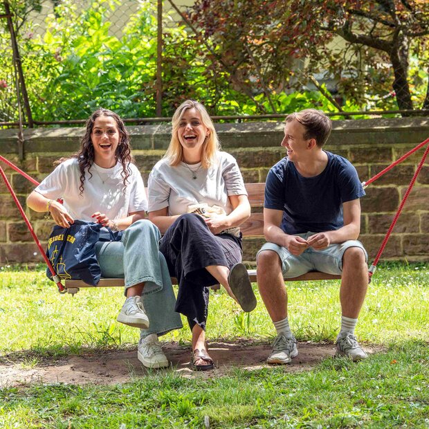Zwei Studentinnen und ein Student sitzen in der Hängeschaukel. Alle drei lachen deutlich. Im Hintergrund sieht man den Innenhof der Hochschule.