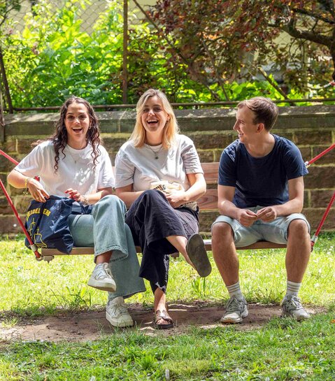 Zwei Studentinnen und ein Student sitzen in der Hängeschaukel. Alle drei lachen deutlich. Im Hintergrund sieht man den Innenhof der Hochschule.