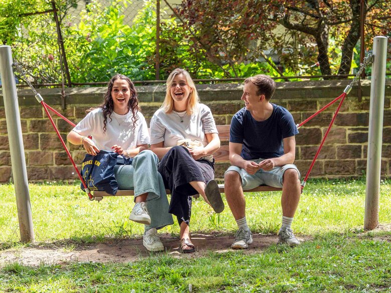 Zwei Studentinnen und ein Student sitzen in der Hängeschaukel. Alle drei lachen deutlich. Im Hintergrund sieht man den Innenhof der Hochschule.
