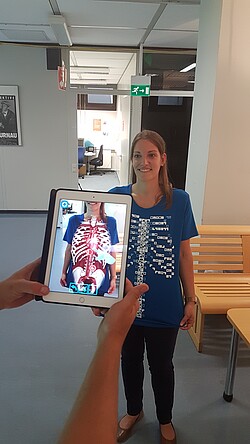 Frau mit einem T-Shirt mit dem knöchernen Brustkorb drauf und ein IPad welches mit einer App vor die rau gehalten wird.