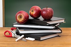 Auf dem Foto sieht man drei Äpfel die auf Büchern auf einem Schreibtisch vor einer Tafel liegen.