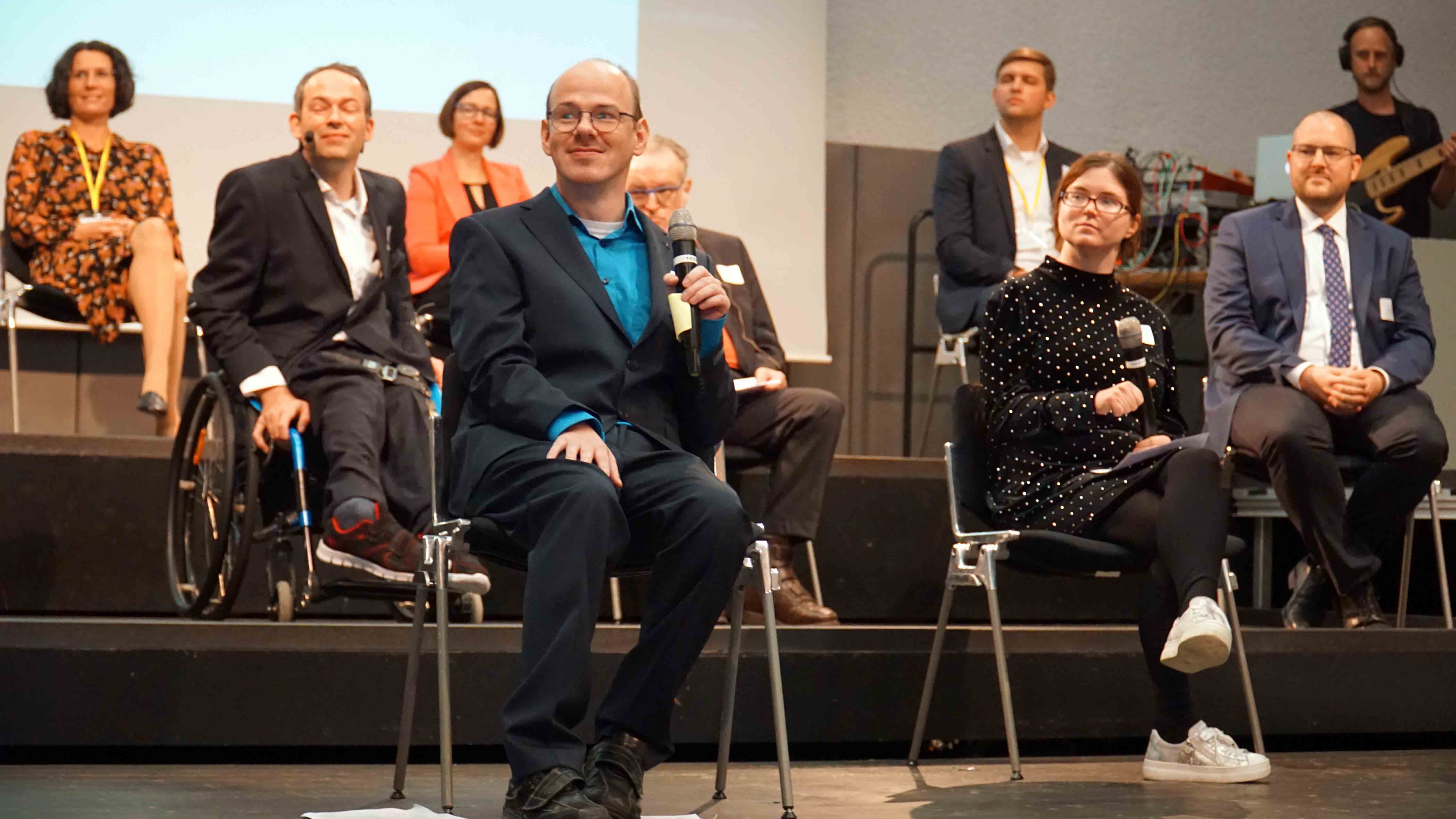 Mehrere Menschen sitzen auf Stühlen oder im Rollstuhl auf der Bühne der Mehrzweckhalle der Pädagogischen Hochschule Heidelberg.