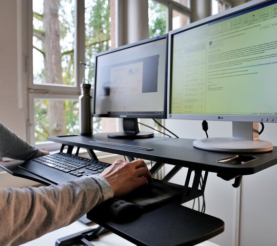 Auf ein Tischaufsteller stehen zwei PC-Bildschirme. Darunter eine Tastatur. Vor dem Aufsteller steht ein Mann, der die Maus bedient.