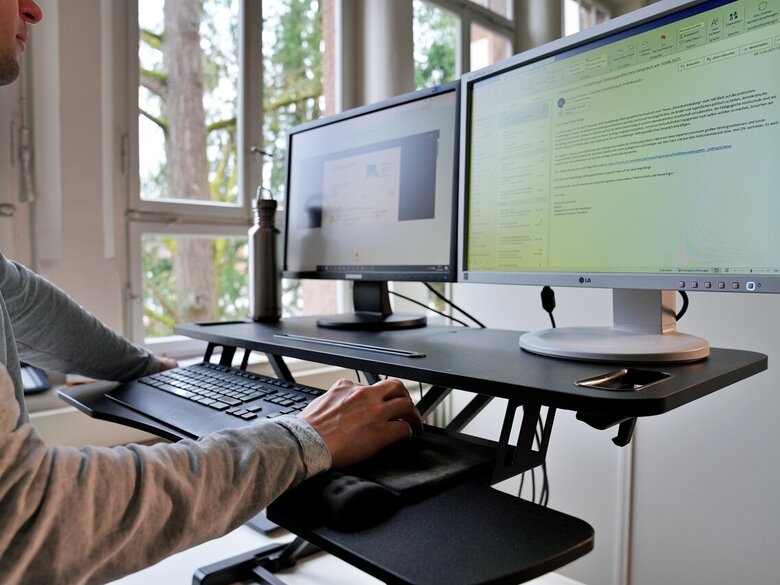 Auf ein Tischaufsteller stehen zwei PC-Bildschirme. Darunter eine Tastatur. Vor dem Aufsteller steht ein Mann, der die Maus bedient.