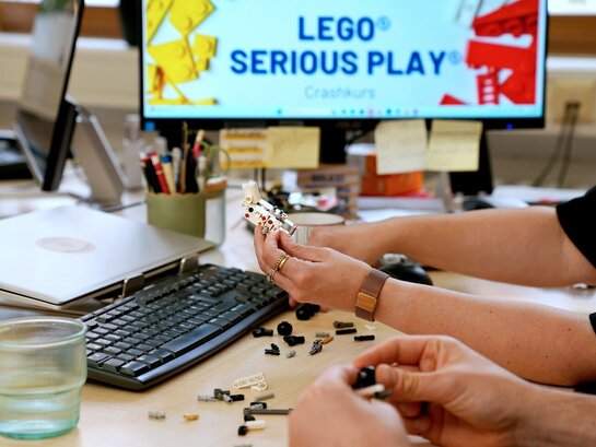 Der Fokus liegt auf den Händern von zwei Personen, die mit Lego etwas bauen. Die Personen sitzen im Büro am Schreibtisch. Auf dem PC-Bildschirm ist "Lego Serious Play" zu lesen.