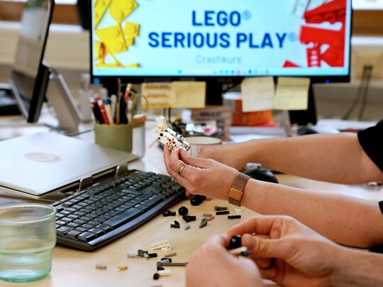 Der Fokus liegt auf den Händern von zwei Personen, die mit Lego etwas bauen. Die Personen sitzen im Büro am Schreibtisch. Auf dem PC-Bildschirm ist "Lego Serious Play" zu lesen.