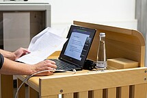 Rednerpult auf dem ein Laptop und eine Flasche Wasser steht. Auf das Rednerpult stützen sich die Arme einer Person, die einen Zettel in der Hand hält, ab.Copyright Pädagogische Hochschule Heidelberg