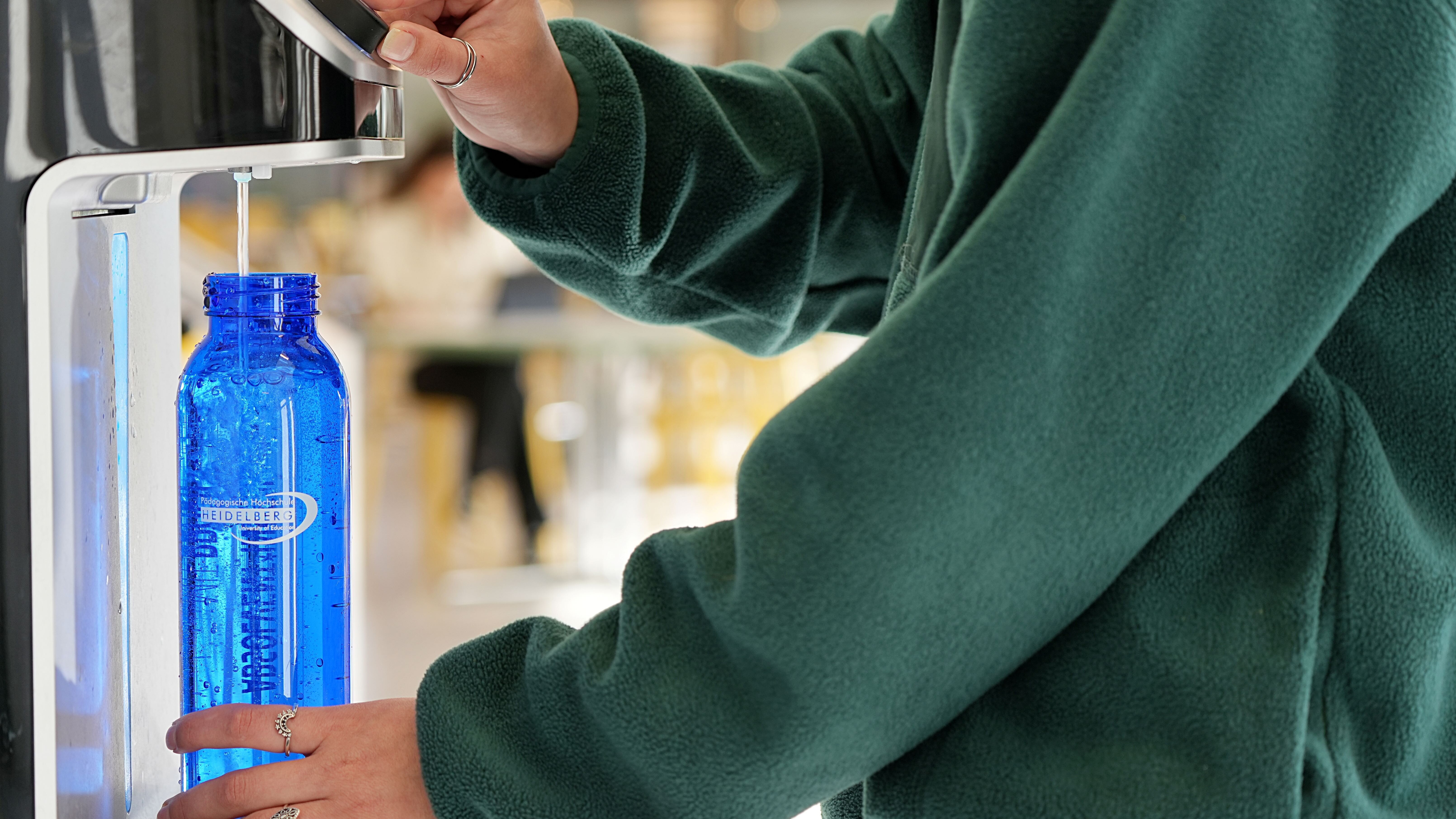 Eine blaue Wasserflasche der PHHD wird in einem Wasserspender mit Sprudelwasser gefüllt