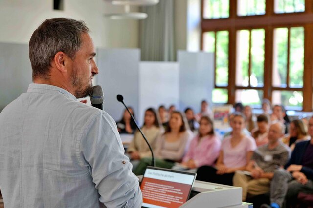 Links im Bild spricht ein Mann in ein Mikro. Man sieht ihn von schräg hinten. Im Hintergrund sieht man Menschen, die in der Aula der Pädagogischen Hochschule Heidelberg sitzen.
