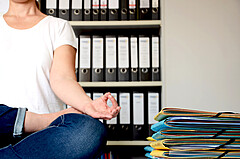 Auf dem Bild ist eine Person zu sehen, die im Meditationssitz auf einem Bürotisch sitzt. Das Gesicht der Person ist nicht zu sehen. Im Hintergrund sieht man unscharf zahlreiche Ordner. Copyright: PH Heidelberg