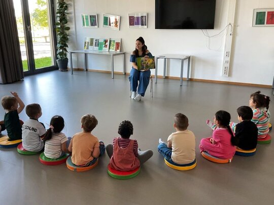 Das Bild zeigt eine Gruppe von Kindern, denen ein Buch vorgelesen wird