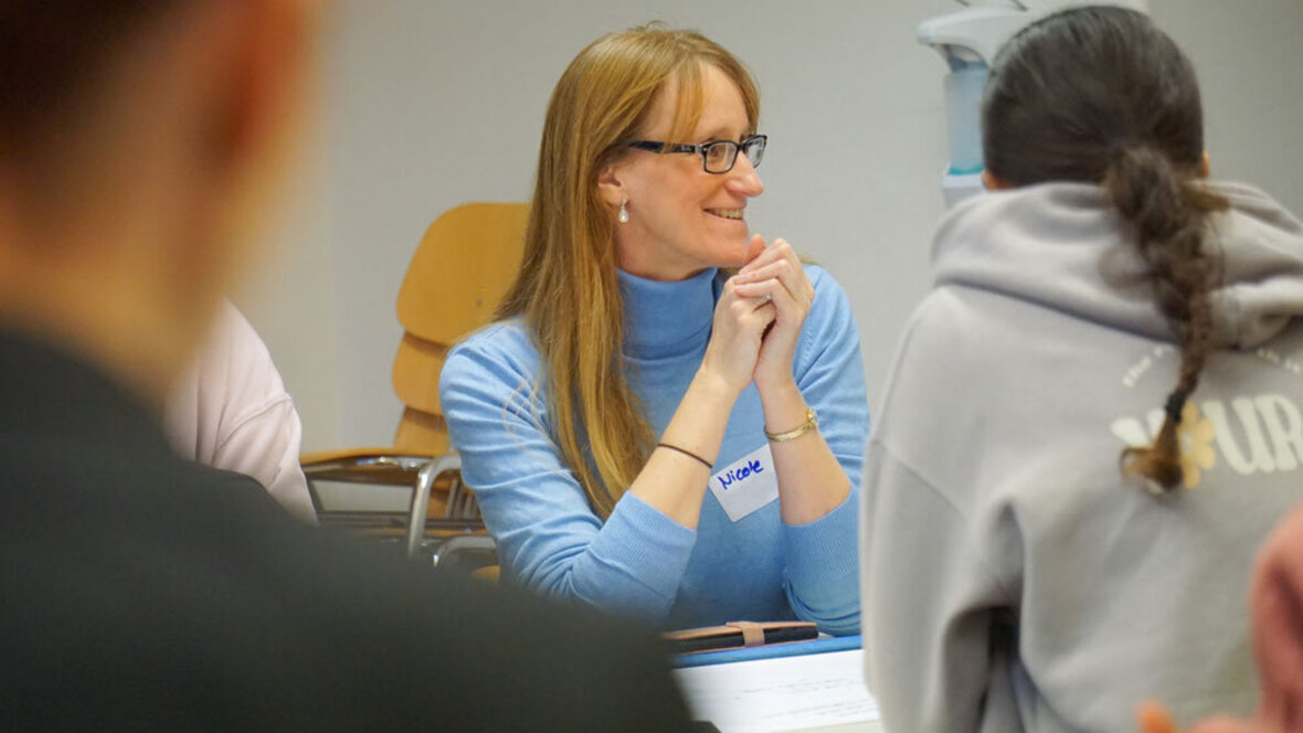 Eine Frau sitzt in einem Workshop und lächelt jemanden an, der außerhalb des Bildes sitzt.