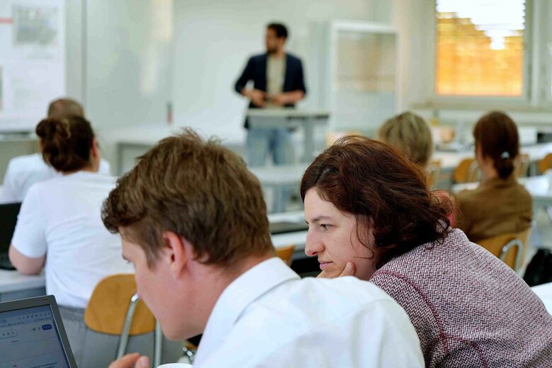 Vorne sieht man einen Doktoranden und eine Doktorandin im Profil. Im Hintergrund unscharf einen Referenten und drei Personen, die ihm zuhören.