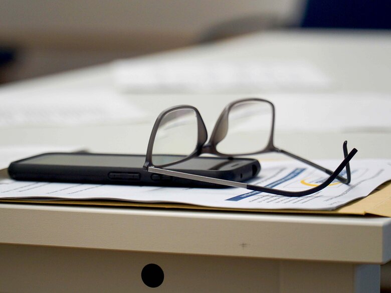 Auf dem Bild sieht man eine Brille und ein Mobiltelefon, die auf einem Tisch liegen. Darunter liegt ein Blatt Papier. 