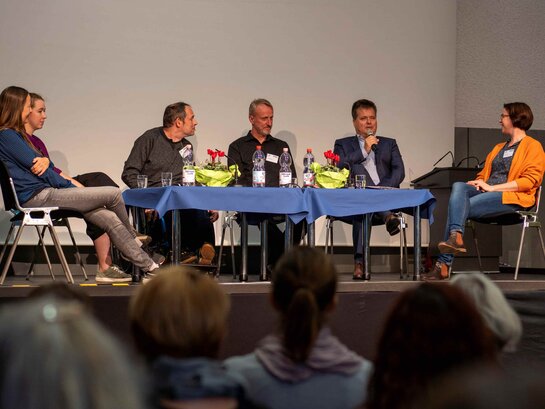 Mehrere Personen sitzen auf einer Bühne um einen Tisch. Sie sind Teil des Podiums der Tagung "Expert:innen in eigener Sache in Forschung, Lehre und beruflicher Bildung"