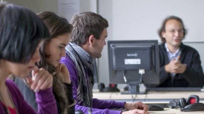 Im Vordergrund lauschen drei Studenten dem Dozenten der im Hintergrund sitzt.