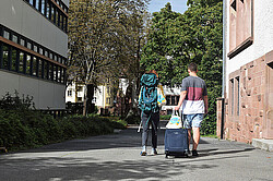 Das Symbolbild zeigt zwei Studierende von hinten. Sie laufen mit Rucksack und Koffer in den Innenhof der Hochschule. Studierende mit Koffern