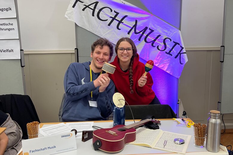 Zwei Studierende der Musikfachschaft sitzen am Studieninformationstag am Stand des Fachs Musik. Eine hält eine Rassel in der Hand, einer eine Ratsche. Auf dem Tisch liegen weitere Instrumente.