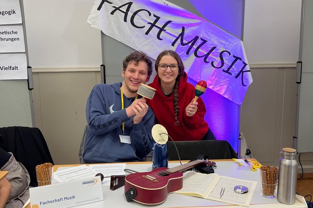 Zwei Studierende der Musikfachschaft sitzen am Studieninformationstag am Stand des Fachs Musik. Eine hält eine Rassel in der Hand, einer eine Ratsche. Auf dem Tisch liegen weitere Instrumente.