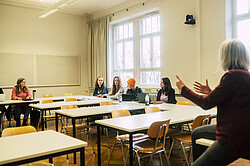 Das Foto zeigt eine Dozentin vor einer Gruppe Studierenden in einem Seminarraum stark gestikulierend.