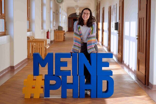 Eine junge Studentin steht hinter den Buchstaben #PHHD in den FLuren des Altbaus.