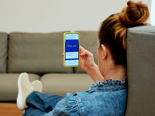 Eine Person sitzt in einem Sessel. Das Foto blickt ihr über die Schulter. Sie hält ein Handy in der Hand, auf dem man den Newsletter der Pädagogischen Hochschule Heidelberg sieht.