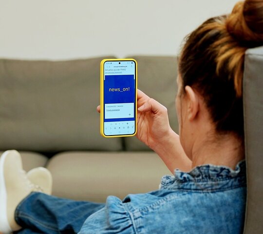 Eine Person sitzt in einem Sessel. Das Foto blickt ihr über die Schulter. Sie hält ein Handy in der Hand, auf dem man den Newsletter der Pädagogischen Hochschule Heidelberg sieht.