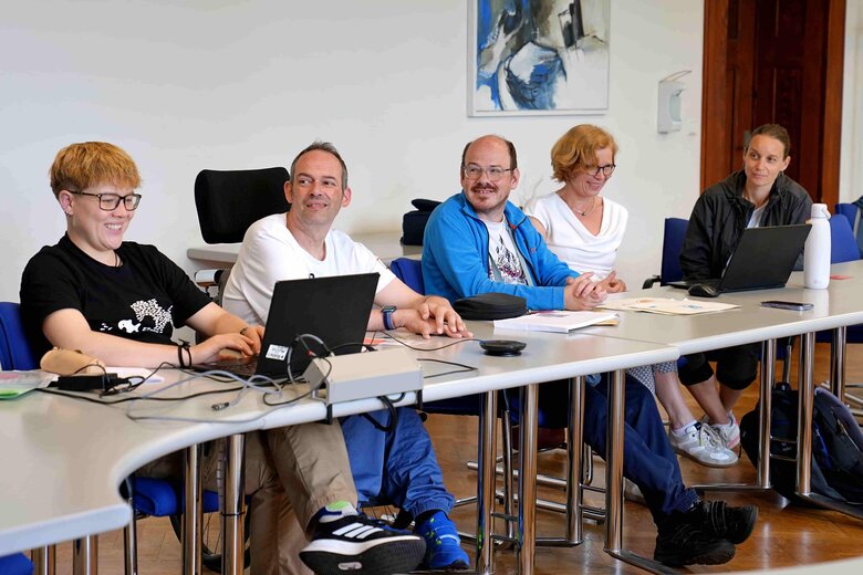 Das Bild zeigt Mitglieder des Forschungsplenums. Sie sitzen an einem Tisch. Eine Person hat einen Laptop vor sich, eine Person sitzt in einem Rollstuhl.