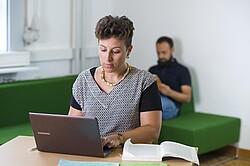 Frau am Laptop und einen Mann im Hintergrund auf dem Sofa.