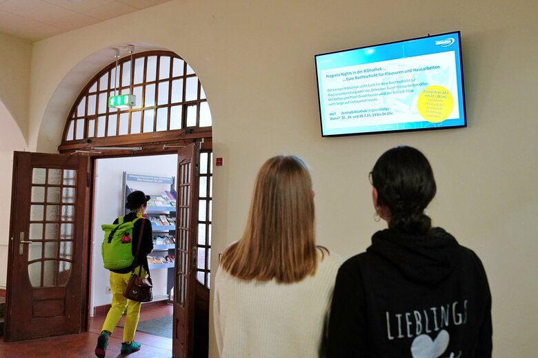 Zwei Frauen schauen auf das digitale Schwarze Brett im Altbau. Man sieht die beiden von hinten. Im Hintergrund läuft eine Person durch die offenen Tür; auch sie ist von hinten zu sehen.