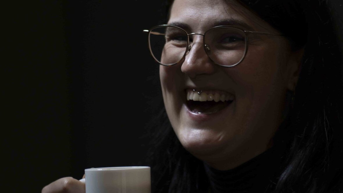 Das Bild zeigt eine Frau, die eine Kaffeetasse in der Hand hält. Die Frau lacht.