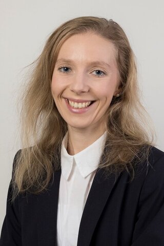 Zu sehen ist ein Portraitfoto der Alumna Jasmin Traum vor einer hellgrauen Leinwand.