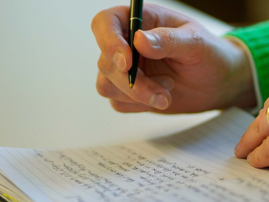 Das Bild zeigt eine Hand, die einen Stift hält und in ein Notizbuch scheibt.