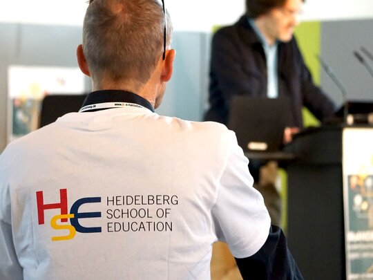 Das Bild zeigt die Rückenansicht eines Mannes in einem T-Shirt, auf dem das Logo der HSE abgebildet ist. Im Hintergrund ist verschwommen ein Referent am Rednerpult zu erkennen.