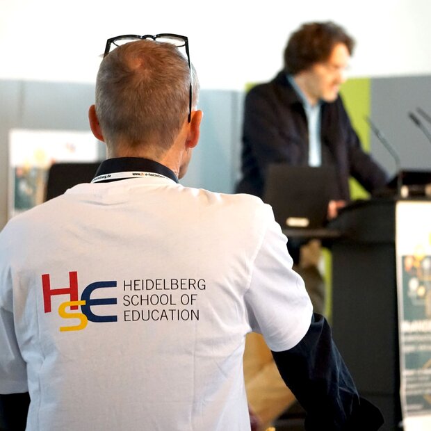 Das Bild zeigt die Rückenansicht eines Mannes in einem T-Shirt, auf dem das Logo der HSE abgebildet ist. Im Hintergrund ist verschwommen ein Referent am Rednerpult zu erkennen.