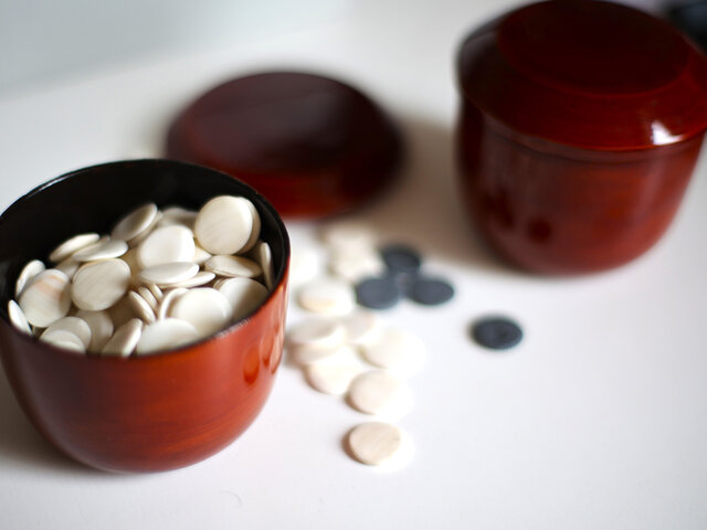 Auf einem Tisch stehen zwei mit weißen und schwarzen, runden Go-Spielsteinen gefüllte rote Becher.