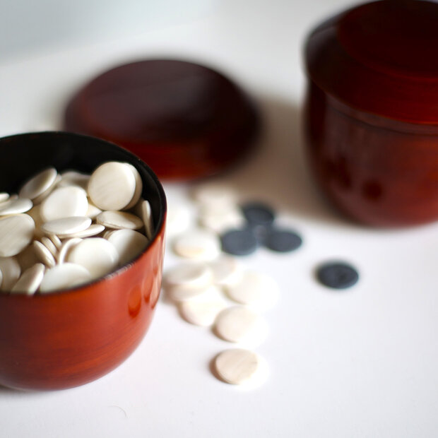 Auf einem Tisch stehen zwei mit weißen und schwarzen, runden Go-Spielsteinen gefüllte rote Becher.