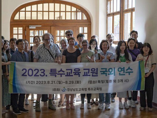 Das Bild zeigt die koreanische Delegation, eine Gruppe von ca. 20 Personen, gemeinsam mit Mitarbeitenden des AW-ZIB. Die Gruppe steht im Flur des Altbaus der PH Heidelberg und hält ein Plakat vor sich, auf dem koreanische Schriftzeichen stehen. 