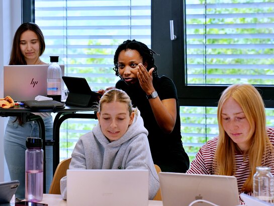 Das Foto zeigt Studierende vor jeweils einem Laptop sitzend oder stehend.