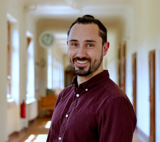 Porträtaufnahme von Philipp Hill im Altbau der Pädagogischen Hochschule Heidelberg. Philipp Hill hat dunkle Haare, trägt ein bordeuxrotes Hemd und lächelt in die Kamera.